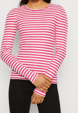 Frau trägt ein langärmeliges rosa-weiß horizontal gestreiftes Oberteil, schwarze Hose, Hände vor der Taille verschränkt.