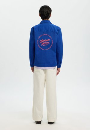 Háttal álló személy, kék dzsekit viselve, amelyen piros "Amour Hotels" felirat található, és fehér, bő szárú nadrágot visel, egyszerű fehér háttér előtt.