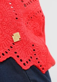 Röd broderad topp med intrikata mönster och utskärningar längs nederkanten, med ett litet guld "Superdry"-märke fäst.