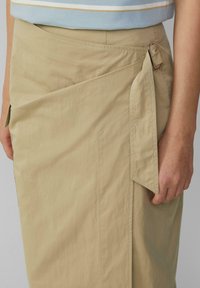 Beige wrap skirt made of lightweight fabric, featuring a side tie, pocket, and visible stitching detail. Smooth texture with a relaxed fit.