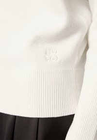 Witte gebreide trui met geribbelde zoom en manchetten; heeft een reliëf "HUGO"-logo; gladde textuur.