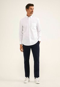 Weißes Hemd mit Knöpfen und Brusttasche, kombiniert mit marineblauen Hosen und weißen Sneakern. Das Hemd hat einen klassischen Kragen und lange Ärmel.