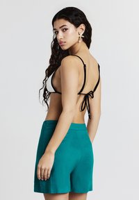 Giovane donna con lunghi capelli ricci, indossa un bikini bianco e nero, pantaloni corti a vita alta turchesi e orecchini a forma di stella marina, mentre guarda sopra la spalla.