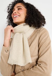 Écharpe tricotée beige avec une texture douce, dotée de subtils éclats. Portée lâchement autour du cou, assortie à un manteau beige.