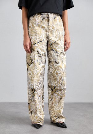 Pantaloni a gamba larga in un tessuto bianco con intricati motivi floreali in oro e nero. I pantaloni hanno una vestibilità rilassata e sono abbinati a scarpe nere con punta.