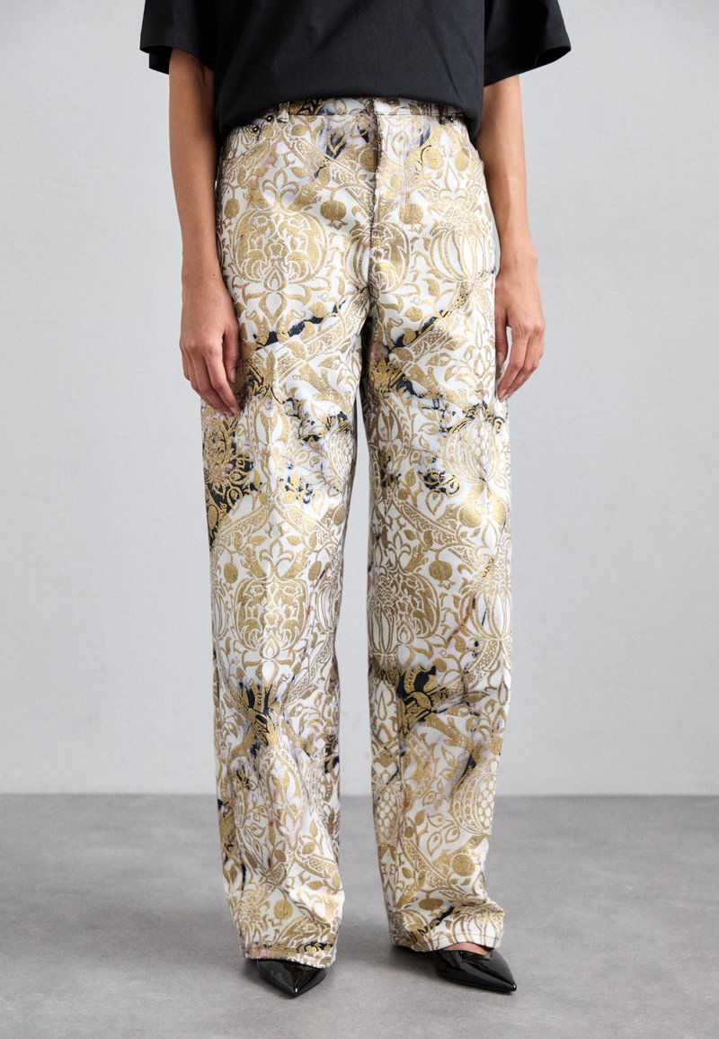 Pantaloni cu croi larg dintr-un material alb, cu modele florale intricate în nuanțe de auriu și negru. Pantalonii au un fit confortabil și sunt purtați cu pantofi negri cu vârf ascuțit.