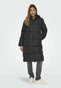 Manteau long matelassé noir avec une capuche, doté d'une fermeture éclair devant, de segments horizontaux et d'une silhouette droite, associé à un pantalon gris et des baskets claires.