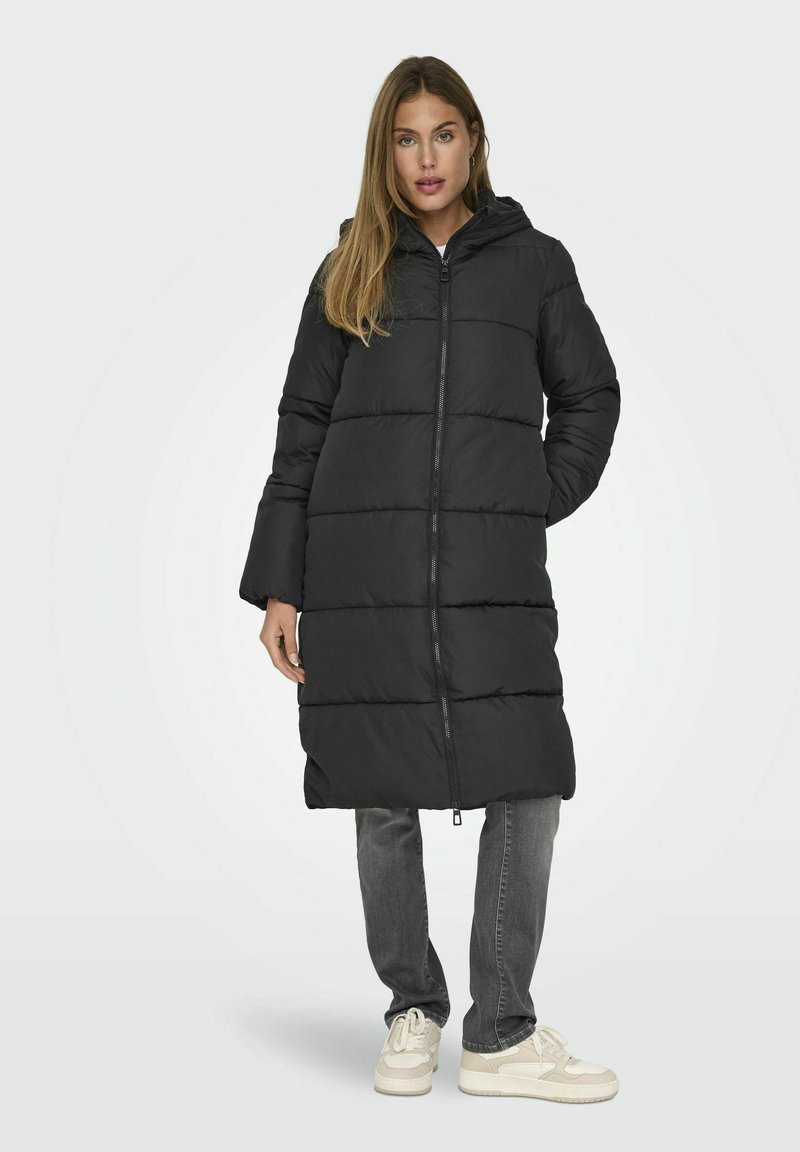 Manteau long matelassé noir avec une capuche, doté d'une fermeture éclair devant, de segments horizontaux et d'une silhouette droite, associé à un pantalon gris et des baskets claires.