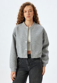 Gekürzter grauer Sweatshirt aus weichem Material mit Rundhalsausschnitt, Druckknopfverschluss und langen Ärmeln. Kombiniert mit schwarzen Jeans.