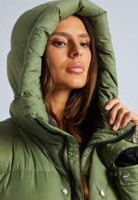 Zaļš pūkainais apģērbs ar lielu kapuci, kas aprīkots ar priekšējo rāvējslēdzēju un pogu aizdari. Audums izskatās gluds ar nelielu spīdumu.