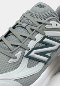 Jasnoszare buty sportowe posiadają siateczkową cholewkę, teksturowane detale, płaskie szare sznurowadła oraz wyraziste turkusowo-białe logo po stronie.