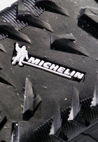 Gros plan sur la bande de roulement noire d'un pneu Michelin avec des rainures profondes et angulaires, ainsi que le logo Michelin présentant la silhouette de Bibendum en blanc.