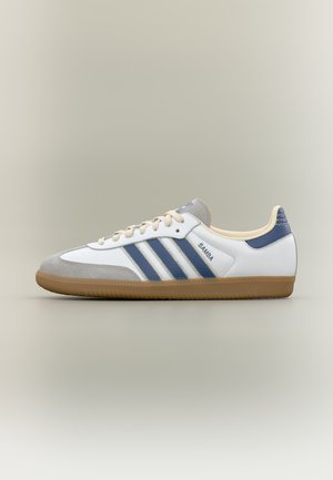Baskets Adidas Samba blanches et grises avec des bandes bleues, des lacets beige et une semelle en gomme sur un fond neutre.
