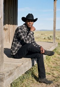 Homme portant un chapeau de cowboy noir, une chemise à carreaux et un pantalon noir assis sur une terrasse en bois à la campagne par une journée ensoleillée.