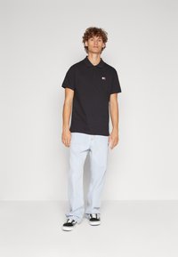 Camisa polo negra con cuello y pequeño logo bordado, combinada con jeans azul claros y zapatillas negras, de pie contra un fondo liso.