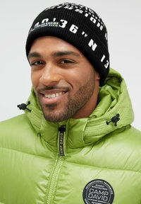 Black knit beanie featuring white text and graphic accents, paired with a shiny green puffer jacket with a round logo patch.