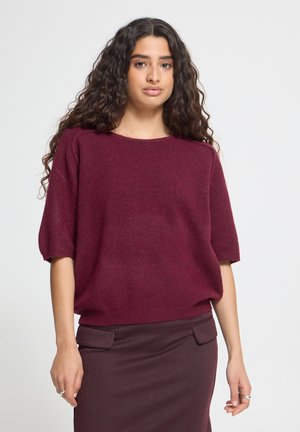 Femme aux longs cheveux bouclés portant un pull bordeaux à manches courtes et une jupe bordeaux foncé avec des rabats de poche, debout devant un fond uni.