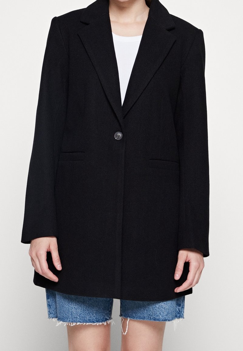 Blazer nero su misura con un unico bottone, rever a punta e due tasche frontali, abbinato a pantaloni corti in denim strappato.
