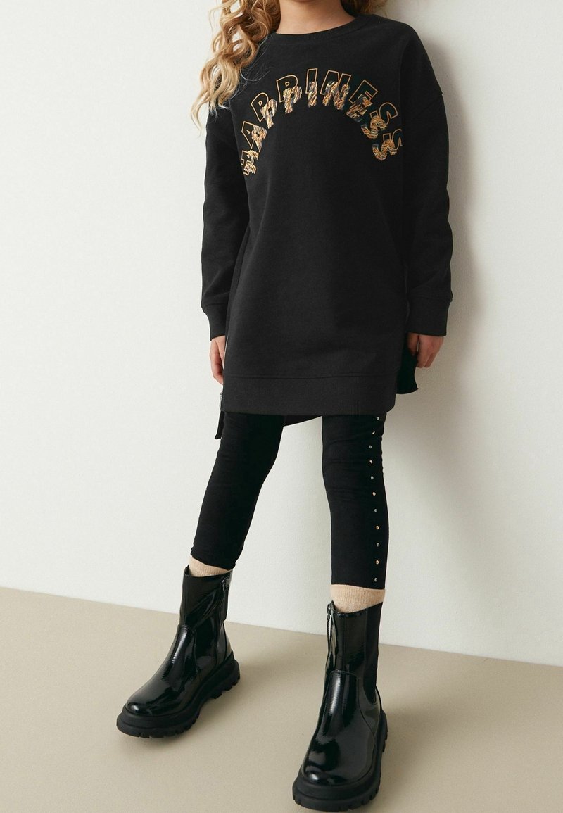 Robe sweat-shirt noire avec le texte « HAPPINESS » en accents dorés, associée à des leggings noirs ornés de clous dorés, et des bottines noires brillantes.