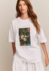 Bijela pamučna oversized majica s kratkim rukavima koja sadrži floralni grafički print u tamnim bojama s natpisom "Revival" i okruglim izrezom.