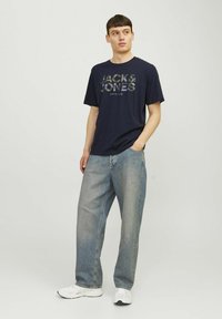 Tricou navy cu guler rotund, decorat cu un grafic de camuflaj "JACK & JONES", asortat cu blugi largi de nuanță deschisă și adidași albi.