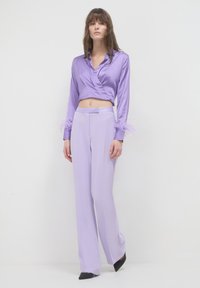 Lila Satin-Wickelbluse mit langen Ärmeln, verziert mit Federn an den Manschetten, kombiniert mit maßgeschneiderten lila Hosen und schwarzen Pumps.