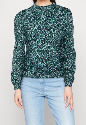 Schwarzes Langarmoberteil mit einem grünen und blauen abstrakten Tiermuster, hohem Kragen und gerafften Details am Saum. Wird mit hellblauen Jeans getragen.