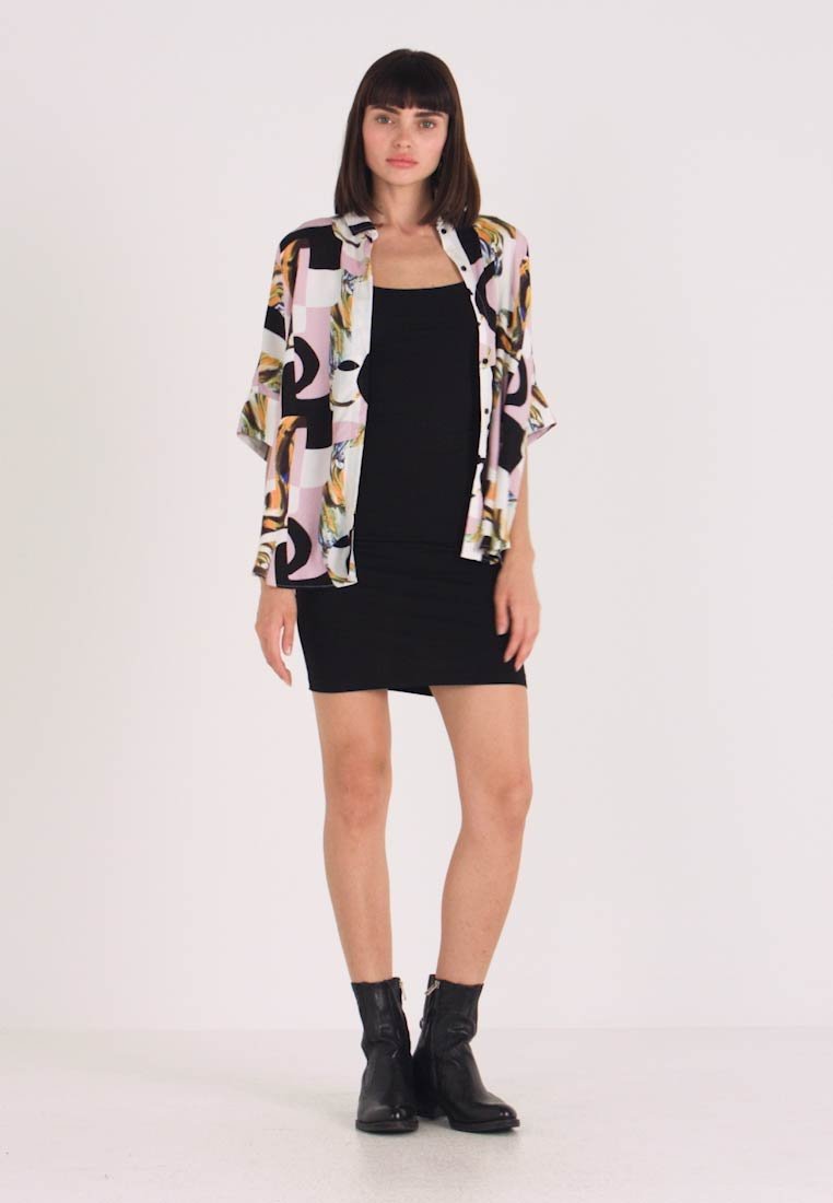 Robe noire ajustée superposée sous une chemise ample à motifs colorés, présentant des designs géométriques et floraux. La mannequin porte des bottines noires.