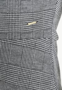 Grått och svart rutigt tyg med varierande texturer och mönster, och en liten guldlogotyp i mitten.