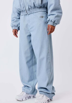 Personne portant un jean baggy bleu clair avec une taille et des poignets élastiques, debout en baskets blanches sur un fond uni.