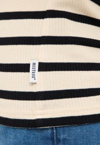 Haut en coton côtelé avec des rayures horizontales noires et crème. Comprend une étiquette en tissu blanc avec "MUSTANG" en bas.