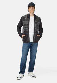 Donkergrijze gebreide cardigan met gepatterneerde accenten, open voorkant, twee voorzakken, gedragen over een witte shirt en gecombineerd met blauwe jeans en witte sneakers.