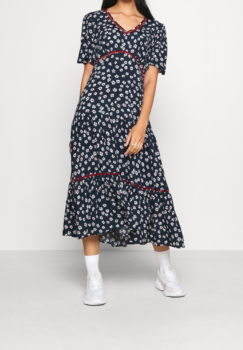 Femme portant une robe midi fleurie bleu marine avec une bordure rouge, des baskets blanches et des chaussettes blanches, debout devant un fond clair uni.
