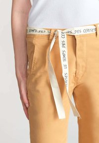 Pantaloni di cotone gialli con una cintura bianca a corda e testo nero. Inquadratura ravvicinata che si concentra sulla vita e sulla parte superiore della gamba, con texture liscia visibile.