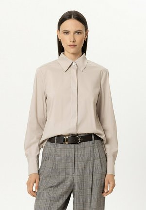 Woman with long dark hair wears beige buttoned shirt tucked into gray plaid pants with black belt, standing against plain background.