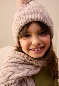 Light pink knitted hat with a pom-pom, paired with a matching cable-knit scarf. The scarf drapes around the neck against a green top.
