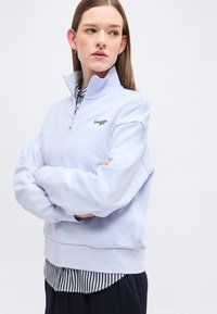 Femme aux longs cheveux portant une veste zippée bleu clair avec un petit logo, une chemise rayée en dessous, et un pantalon foncé, regardant sur le côté.