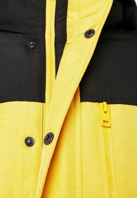 Żółto-czarna pikowana kurtka z zamkiem błyskawicznym, z czarnymi guzikami, gładką teksturą tkaniny i widocznymi detalami przeszycia.