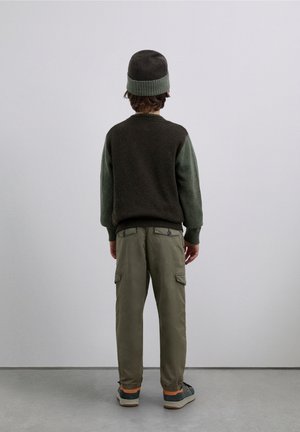 Maglione marrone e verde lavorato a maglia, pantaloni cargo e berretto a maglia; caratterizzati da polsini a coste, tasche e silhouette rilassata.