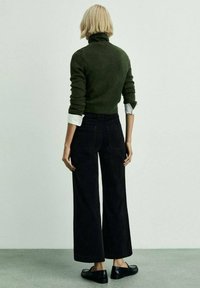 Maglione turtleneck verde con polsini risvoltati, abbinato a pantaloni neri a vita alta e a gamba larga. Le scarpe nere completano il look.