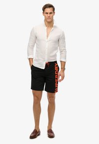 Pantaloni da nuoto neri con un logo arancione "SUPERDRY" di lato, abbinati a una camicia bianca in lino e mocassini marroni. Vita con lacci.