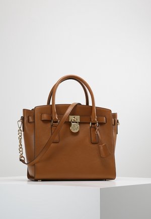 Brązowa skórzana torebka z podwójnymi uchwytami, odpinanym paskiem na ramię oraz złotym zamkiem i detalem łańcuszka na białym postumencie.