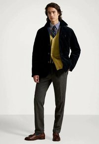 Jeune homme portant une veste matelassée noire, un cardigan jaune moutarde, une chemise rayée avec une cravate, un pantalon gris et des mocassins bruns, debout avec une main dans sa poche.