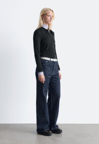 Black long-sleeve button-up shirt paired with dark blue wide-leg jeans, white belt, and chunky black shoes on a neutral background.