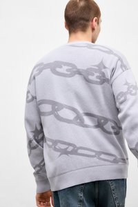 Mand iført en lysepurpur trøje med et mørkepurpur kædemønster, vendt væk, kombineret med blå jeans mod en enkel hvid baggrund.