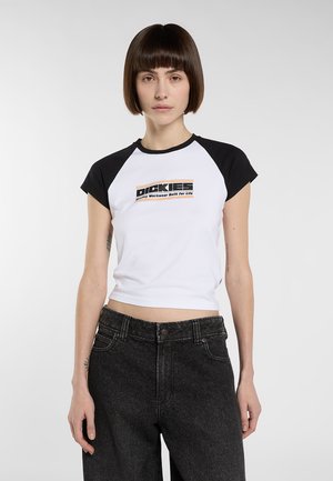 Mujer con el pelo corto y castaño que lleva una camiseta corta Dickies blanca y negra y unos vaqueros negros de talle alto, de pie frente a un fondo simple.
