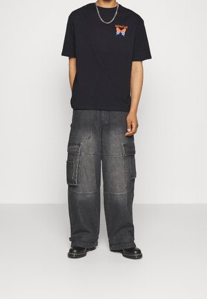 Persona con camiseta gráfica negra, pantalones cargo anchos de color gris oscuro con múltiples bolsillos, zapatos negros y collar de cadena plateada.