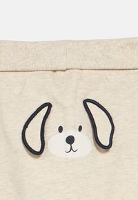 Το ανοιχτό μπεζ ύφασμα διαθέτει το πρόσωπο ενός καρτούν σκύλου με μαύρες κεντητές λεπτομέρειες για τα μάτια και τα αυτιά, δημιουργώντας ένα παιχνιδιάρικο σχέδιο στη μέση.