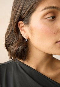 Pendientes de aro de plata con un diseño floral que presenta una piedra preciosa central, colgando justo debajo del lóbulo de la oreja. El fondo es neutro.