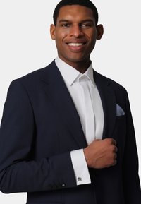 Navy tailored suit jacket with a white dress shirt, featuring a pocket square, silver cufflink, and a textured fabric.